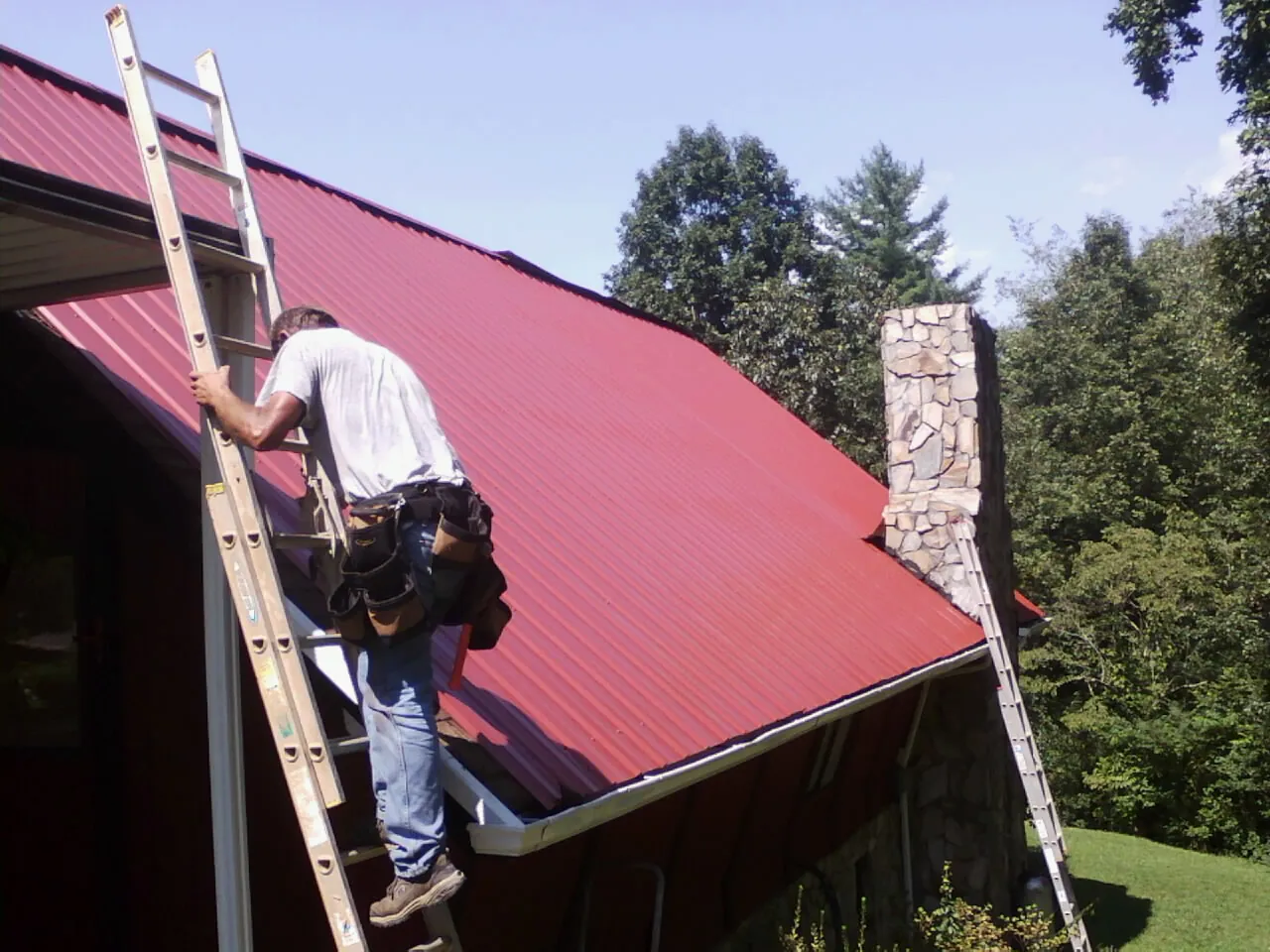 Roofer on a ladder beside a red metal roof and stone chimney — Soffit & Fascia Repair by Roofing Brothers in Bremo Bluff